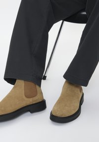 Asmuo sėdi kėdėje, vilkėdamas juodus kelnes ir smėlio spalvos zomšinius Chelsea batus su juodais padais ant baltų grindų.