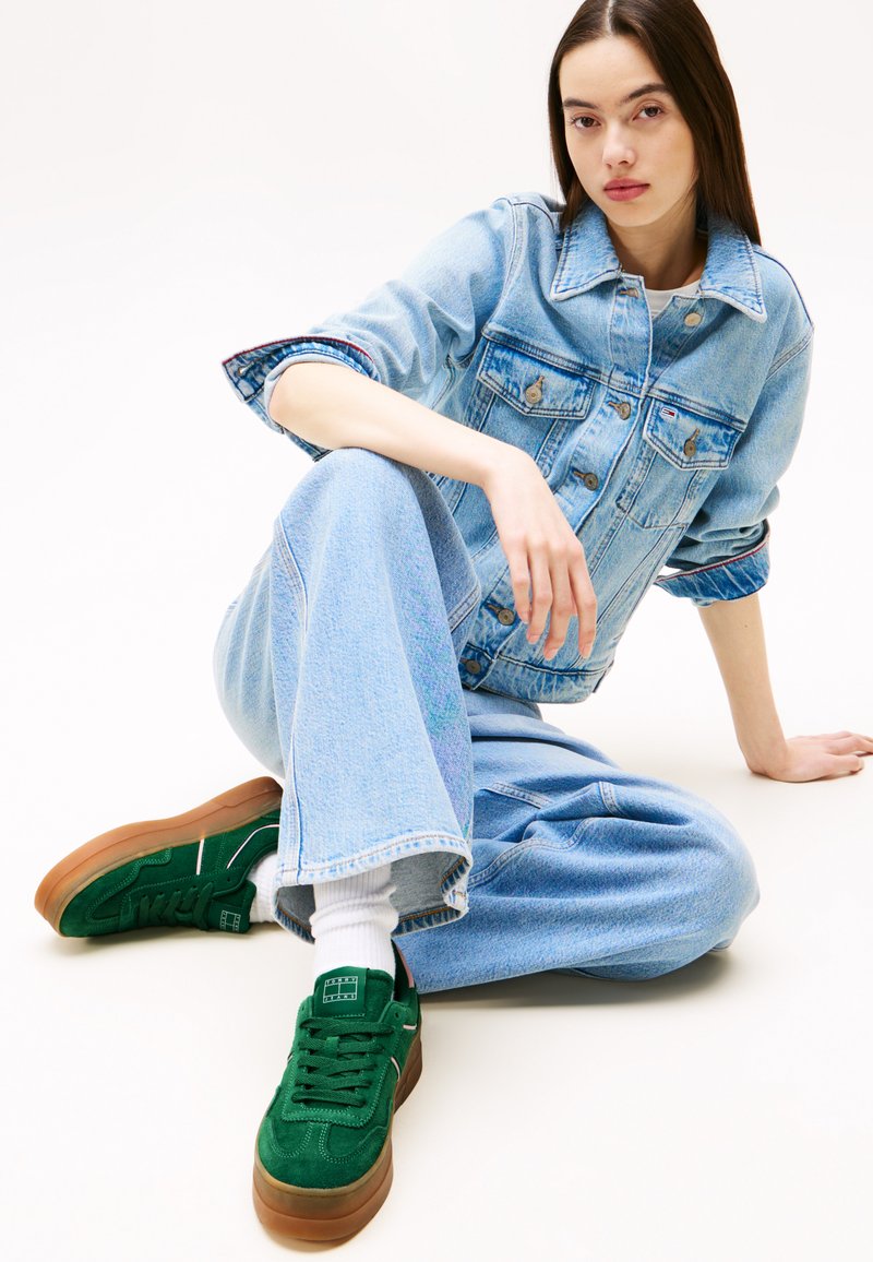 Denimjack en high-waisted jeans in lichtblauw, gecombineerd met groene suède sneakers en witte sokken; model zit tegen een neutrale achtergrond.
