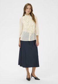 Ivoorwitte blouse met ruches aan de voorkant, gecombineerd met een lange navy rok en zwarte pumps met spits toelopende neus, wat samen een klassieke outfit creëert.