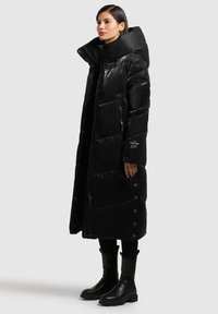 Zwarte, lange donsjas met een hoge kraag, glanzende textuur, drukknoop- en ritssluitingen, zijzakken en een opvallend doorgestikt ontwerp.