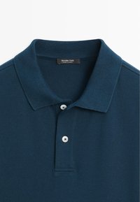 Dunkelblaues, strukturiertes Poloshirt mit zwei weißen Knöpfen und einem umgeschlagenen Kragen, mit dem Etikett Massimo Dutti, hergestellt in der Türkei.