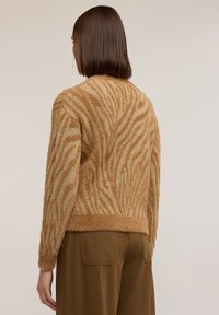 Nainen, jolla on suorat ruskeat hiukset ja joka katsoo poispäin, on pukeutunut pehmeään vaaleanruskeaan neulepaitaan, jossa on teksturoitu seeprahye.