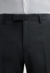 Schwarze Anzughose mit einem dezenten gepunkteten Muster, flachem Vorderteil und Gürtelschlaufen. Glatte Textur, maßgeschneiderte Passform und seitliche Taschen.