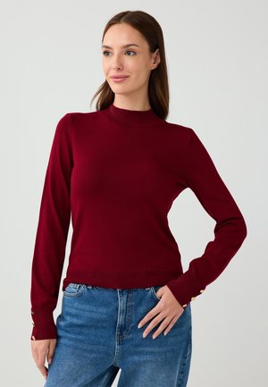 Maglione a maglia bordeaux con collo alto, design slim-fit, maniche lunghe e dettagli in oro sui polsini, abbinato a jeans in denim blu.