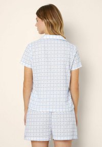 Kvinna med axellångt hår som bär en matchande vit kortärmad topp och shorts med små blå geometriska mönster, vänd bort.