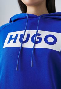 Gros plan d'une personne portant un sweat à capuche bleu avec un logo rectangulaire blanc indiquant "HUGO" sur la poitrine.