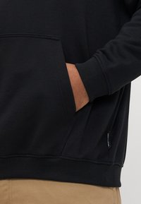 Mână parțial introdusă în buzunarul din față al unui hanorac negru purtat cu pantaloni bej, arătând detalii cu manșete și tiv ribbate.