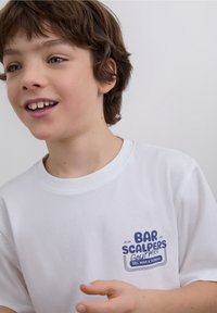 White cotton t-shirt featuring blue graphic text on the left chest area, smooth texture, and a round neckline.