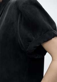 Μαύρο ρούχο με κοντό μανίκι φουσκωτό φορεμένο στο χέρι μπροστά σε λευκό φόντο, δείχνοντας την υφή του υφάσματος και το λεπτομερές γυρισμένο μανίκι.