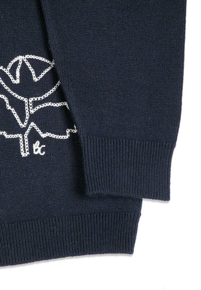 Manche et ourlet d'un pull en maille bleu marine avec un motif de feuille brodé en blanc et de petites lettres "bc" près du poignet.