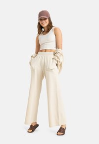 Beige weite Hosen mit Seitentaschen, kombiniert mit einem weißen Cropped-Top und einer mauvefarbenen Mütze. Glatte Stofftextur und lockere Passform.
