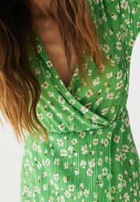 Femme portant une robe verte avec un motif floral blanc et un décolleté en V, avec de longs cheveux bruns partiellement visibles.