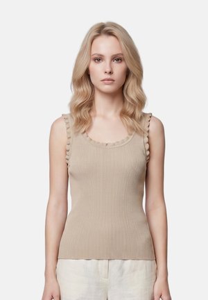 Giovane donna con capelli biondi ondulati, che indossa un top beige senza maniche a coste con bordi arricciati e pantaloni chiari, in piedi, rivolta in avanti.
