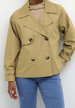 Persona con una chaqueta beige de doble botonadura con botones negros, suéter de cuello alto beige y jeans azules, de pie con una mano en la cadera.