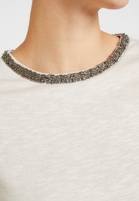 Haut beige clair avec un col texturé orné de perles métalliques et de détails complexes. Le tissu semble doux et léger.