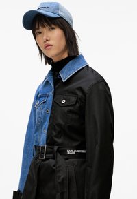 Kurtka z denimu i czarnego materiału o strukturalnym fasonie, z zapięciem na guziki, kieszenią na piersi i paskiem. Uzupełniona niebieską czapką.