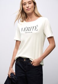 Hvid t-shirt med korte ærmer og sort trykt tekst "VÉRITÉ club de la rue," parret med mørk denimjeans. Glat stof, afslappet pasform.