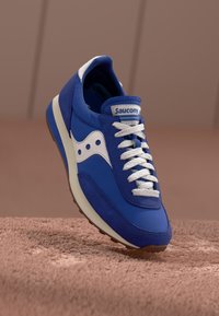 Mėlynas sportinis batelis su zomšos ir odos medžiagomis, baltomis detalėmis, tekstūruotomis raišteliais ir kreiva pada. Ant liežuvio yra "Saucony" ženklas.