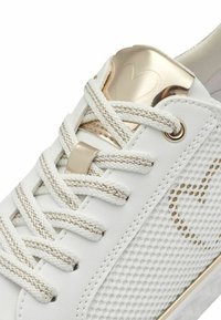 Sneaker bianca con materiali in rete e pelle, dettagli dorati, suola textured e lacci piatti. Presenta un design a cuore metallico sulla linguetta.