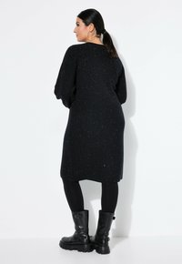 Schwarzes, glitzerndes Kleid mit lockerer Passform, langen Ärmeln und gerundetem Saum. Kombiniert mit schwarzen Leggings und klobigen schwarzen Stiefeln.