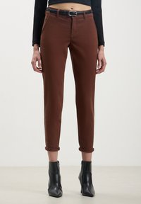 Pantaloni di cotone marroni con una vestibilità slim, orli arrotolati e una cintura nera. Abbinati a stivaletti neri con punta affusolata.