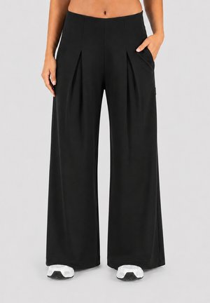Personne portant un pantalon noir taille haute à jambes larges avec des plis devant, mains détendues et baskets blanches sur un fond clair uni.
