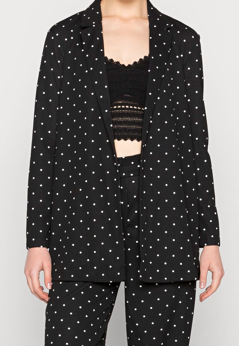 Costume noir à pois blancs porté sur un crop top en dentelle noire, montrant la tenue du cou aux cuisses supérieures.