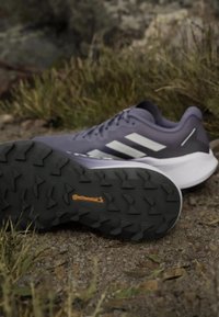 Zapatilla deportiva morada con franjas blancas, suela exterior negra texturizada y un logotipo sutil. Diseño de perfil bajo con un cuello y lengüeta acolchados.