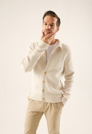 Cárdigan crema con un tejido texturizado, presenta un cierre delantero con botones y un cuello con solapa. Llevado sobre una camisa blanca, combinado con pantalones claros.