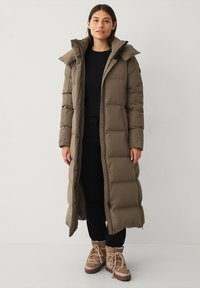 Lange, gevoerde pufferjas in olijfgroen met een capuchon, drukknoopsluitingen en zijzakken, gecombineerd met zwarte broek en bruine veterschoenen.