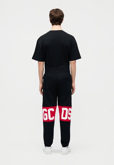 Άνδρας που φοράει μαύρο μπλουζάκι και μαύρη φόρμα με κόκκινες ρίγες και λευκά γράμματα "GCDS" στο πίσω μέρος των ποδιών, στέκεται με την πλάτη στραμμένη.
