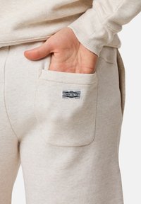 Beige sweatpants har ett texturerat tyg med en bakficka som har en etikett med texten "Original Denim Factory."