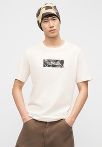 Un jeune homme portant une chemise crème à manches courtes avec un motif rectangulaire, un pantalon marron, les mains dans les poches, et un bonnet à motifs.