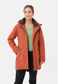 Giacca arancione impermeabile con cappuccio viola scuro, tasche frontali e zip. Indossata sopra una maglietta verde e pantaloni neri.