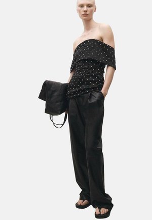 Black off-the-shoulder top with white polka dots, paired with loose-fitting black trousers. Smooth texture, minimalistic design, casual sandals.