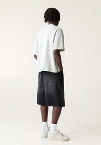 T-shirt in cotone grigio, vestibilità rilassata, maniche corte, abbinato a shorts in denim neri con tasche. Il modello indossa calze bianche e sneakers.
