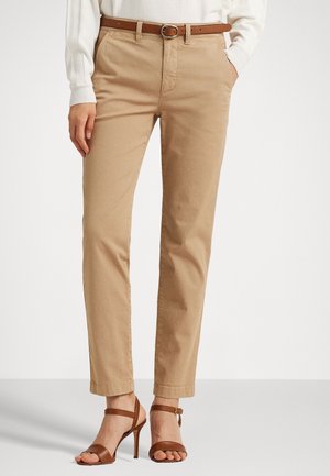 Beige gerade geschnittene Hose mit braunem Gürtel, kombiniert mit braunen, hochhackigen Sandalen mit offenen Zehen und weißem Langarmoberteil. Hände in den Taschen.