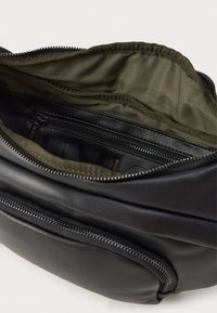 Bolsa de couro preta com fecho éclair aberta para mostrar o revestimento em tecido verde-oliva e um bolso interno com fecho éclair.
