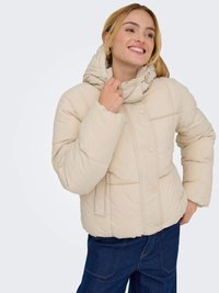 Casaco acolchoado bege com gola alta, fecho de botão e capuz elástico. Apresenta costura em quilt e bolsos laterais. Combinado com jeans azuis.