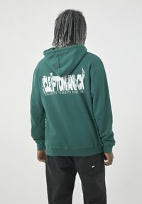 Green hoodie with a drawstring and kangaroo pocket, featuring a large white graphic text on the back. Paired with black pants.