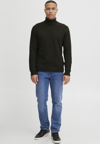 Mørkegrønn, teksturert genser med høy hals og lange ermer, satt sammen med lyse blå jeans og svarte joggesko. Enkel og uformell antrekk.