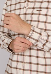 Chemise à boutons avec un motif à carreaux beige et marron, dotée d'un col, de manches longues et d'un petit logo marron au-dessus de la poche.