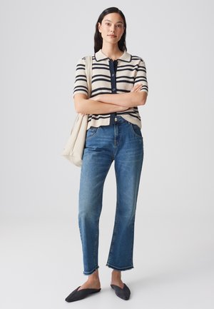 Vrouw staat met gekruiste armen, draagt een gestreept T-shirt met korte mouwen, blauwe jeans, zwarte instapschoenen en draagt een beige schoudertas.