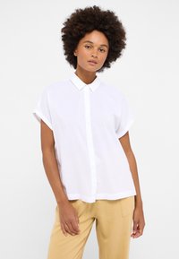 Camisa blanca de manga corta y botones con cuello, hecha de tejido ligero, con un dobladillo recto y mangas enrolladas.