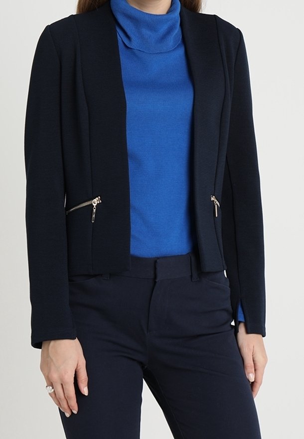 Femme portant un pull à col roulé bleu, un blazer noir ajusté avec des poches zippées et un pantalon noir, debout devant un fond uni.