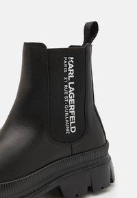 Botín Chelsea negro con material liso y suela de goma texturizada. Presenta un panel elástico lateral y una banda con texto blanco: "KARL LAGERFELD."