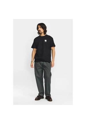 Mann steht, trägt ein schwarzes T-Shirt mit kurzen Ärmeln und kleinem weißem Logo, dunkelgraue Hose und dunkelbraune Schuhe vor weißem Hintergrund.