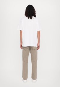 Camiseta blanca de manga corta combinada con pantalones rectos beige y zapatillas blancas, vista desde atrás sobre un fondo liso.