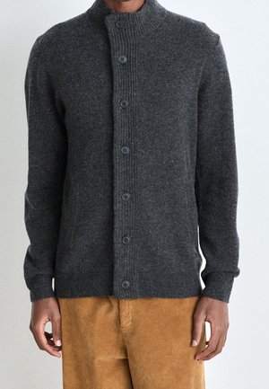 Mężczyzna ubrany w ciemnoszary sweter na guziki i jasnobrązowe spodnie sztruksowe, stojący z ramionami swobodnie opuszczonymi wzdłuż ciała.