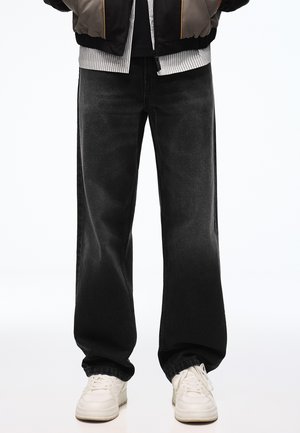 Unterkörper einer Person, die lockere schwarze Jeans, weiße Sneakers, ein gestreiftes Hemd und eine Bomberjacke trägt, mit den Händen in den Taschen.